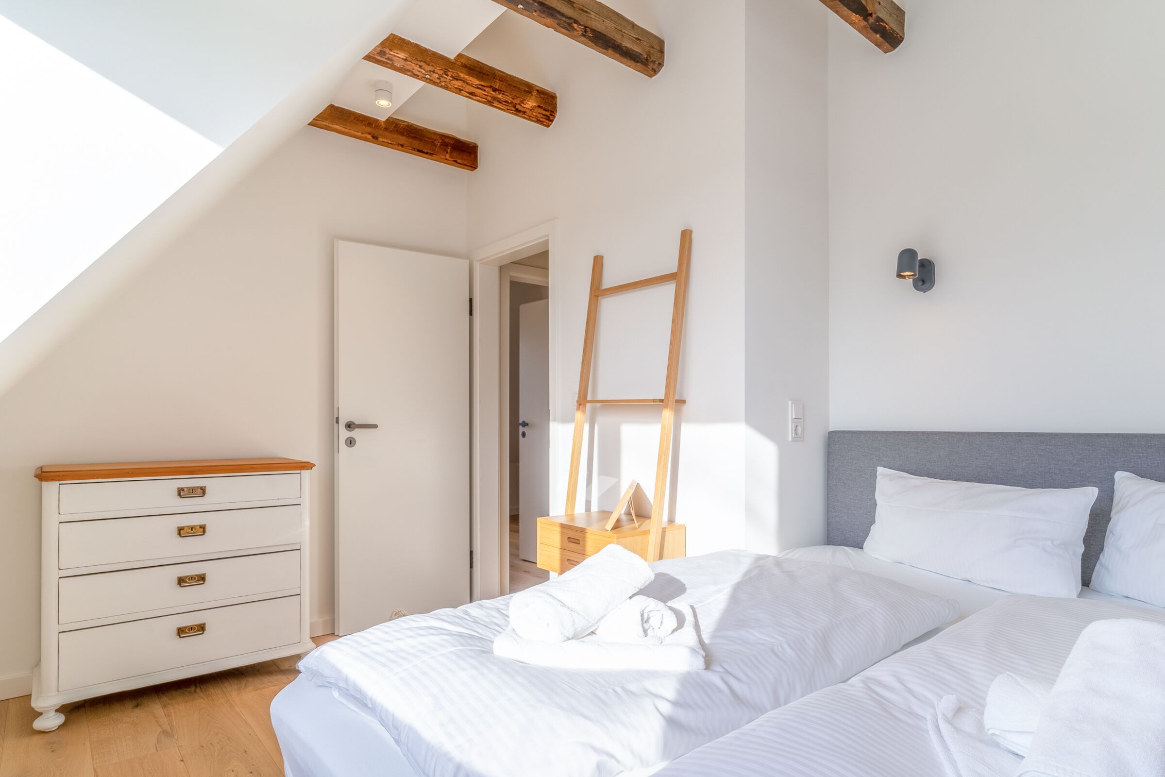 A bedroom with a loft-style ceiling, a wooden dresser, a bed with white linens, and a ladder leaning against the wall.