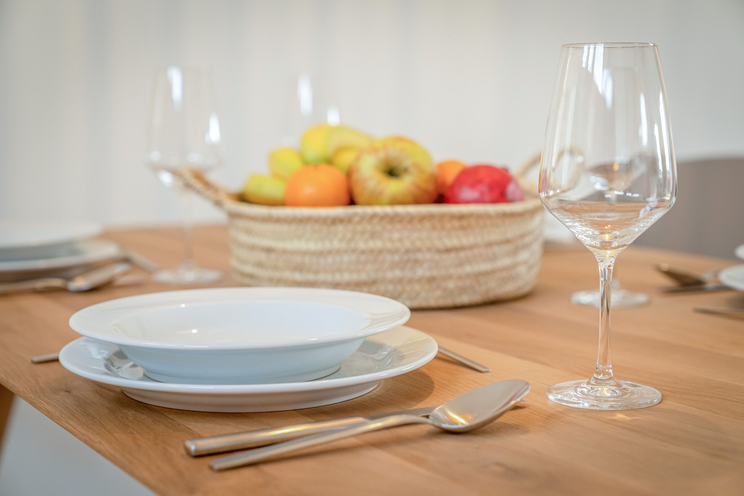 Une table à manger avec un saladier blanc, un verre à vin et un panier de fruits.