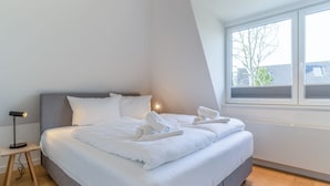 Une chambre moderne avec un grand lit, une table de chevet avec une lampe et une fenêtre donnant sur des immeubles.