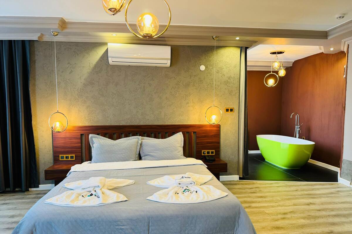 A hotel room with a large bed, a wooden headboard, two bedside tables, a wall-mounted air conditioner, and a green bathtub.
