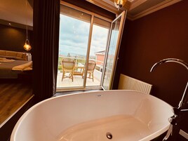 Un baño moderno con una bañera grande, una puerta de cristal que da a un balcón con mobiliario de mimbre y vistas al mar.