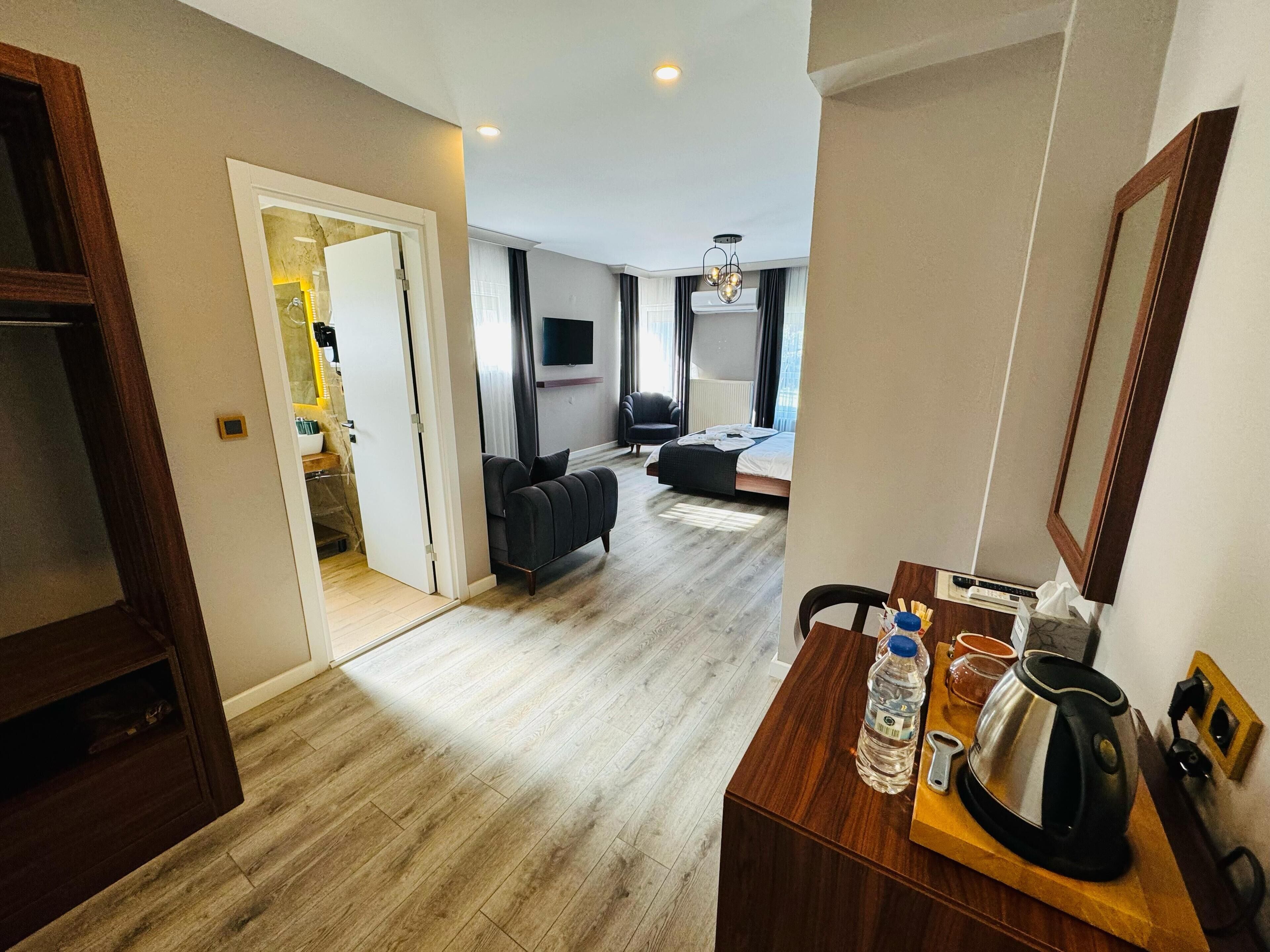 A modern hotel room with a wooden floor, a sofa, a bed, a TV, and a desk with a kettle and water bottle.