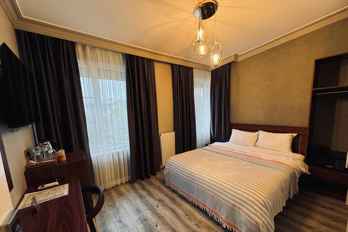 A hotel room with a wooden floor, a bed with a striped blanket, a desk with a chair, a TV, and a window with curtains.