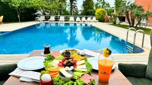 Disposição de refeições junto à piscina, com diversos alimentos e bebidas, incluindo uma salada, frutas e uma bebida com o nome 'Trekkos Suite'.