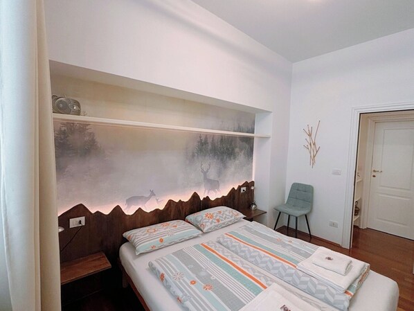 Un dormitorio con cama, una silla, un estante con una obra de arte de temática natural y una puerta.