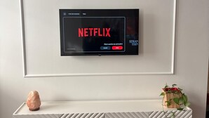 Una sala moderna con un televisor montado que muestra la interfaz de Netflix, un mueble bajo blanco con diseños geométricos y una planta pequeña sobre el mueble.