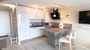 Ett modernt kök med vita skåp, en svart köksfläkt, en inbyggd köksö med barstolar och en platt-tv som är monterad på väggen.