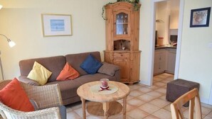 A living room with a brown sofa, a round wooden coffee table, wicker chairs, a wooden cabinet, and a kitchen visible through an open doorway. - App. Stadtoase, Haus Nordland (Sylt)