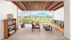 A spacious room with a high ceiling, wooden beams, a large window offering a scenic view, a bookshelf, a seating area with a sofa and armchairs, and a small table with a teapot. - Villa Chrysallis (Apokoronas)
