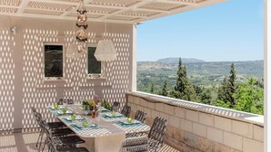 An outdoor dining area with a view of the countryside, featuring a long table set with chairs and a pergola overhead. - Villa Chrysallis (Apokoronas)