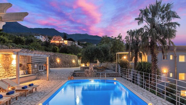 A swimming pool with lounge chairs and a stone wall, surrounded by a stone deck and palm trees, with a mountainous backdrop. - Villa Chrysallis (Apokoronas)