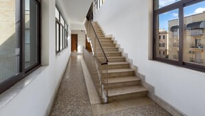 A corridor with a staircase leading to an upper floor, large windows on both sides, and a view of neighboring buildings.