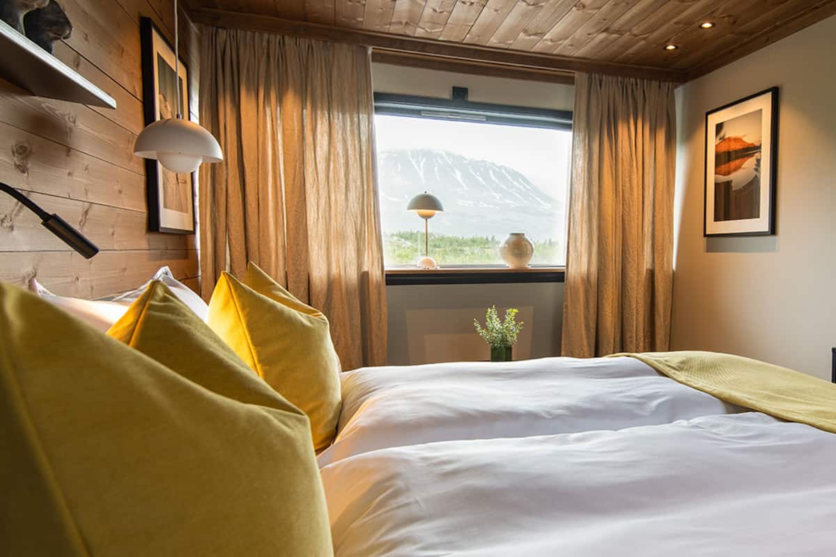 A bedroom with a wooden ceiling, a large bed with white bedding, yellow pillows, a window with a view of a mountain, and a vase with flowers on the windowsill.