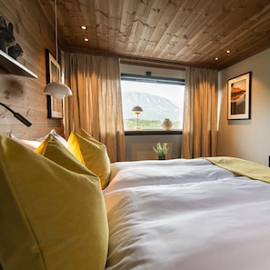 Une chambre avec un plafond en bois, un grand lit avec une literie blanche, des coussins jaunes, une fenêtre donnant sur une montagne et un vase rempli de fleurs posé sur le rebord de la fenêtre.