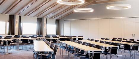 Un salón de conferencias con varias mesas y sillas, luces circulares en el techo y un alto techo con vigas de madera.