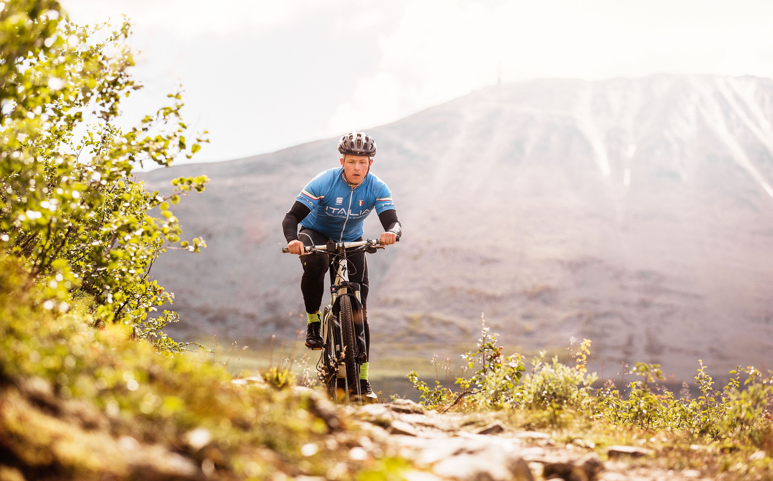 A mountain biker on a trail with greenery and a mountain in the background.
