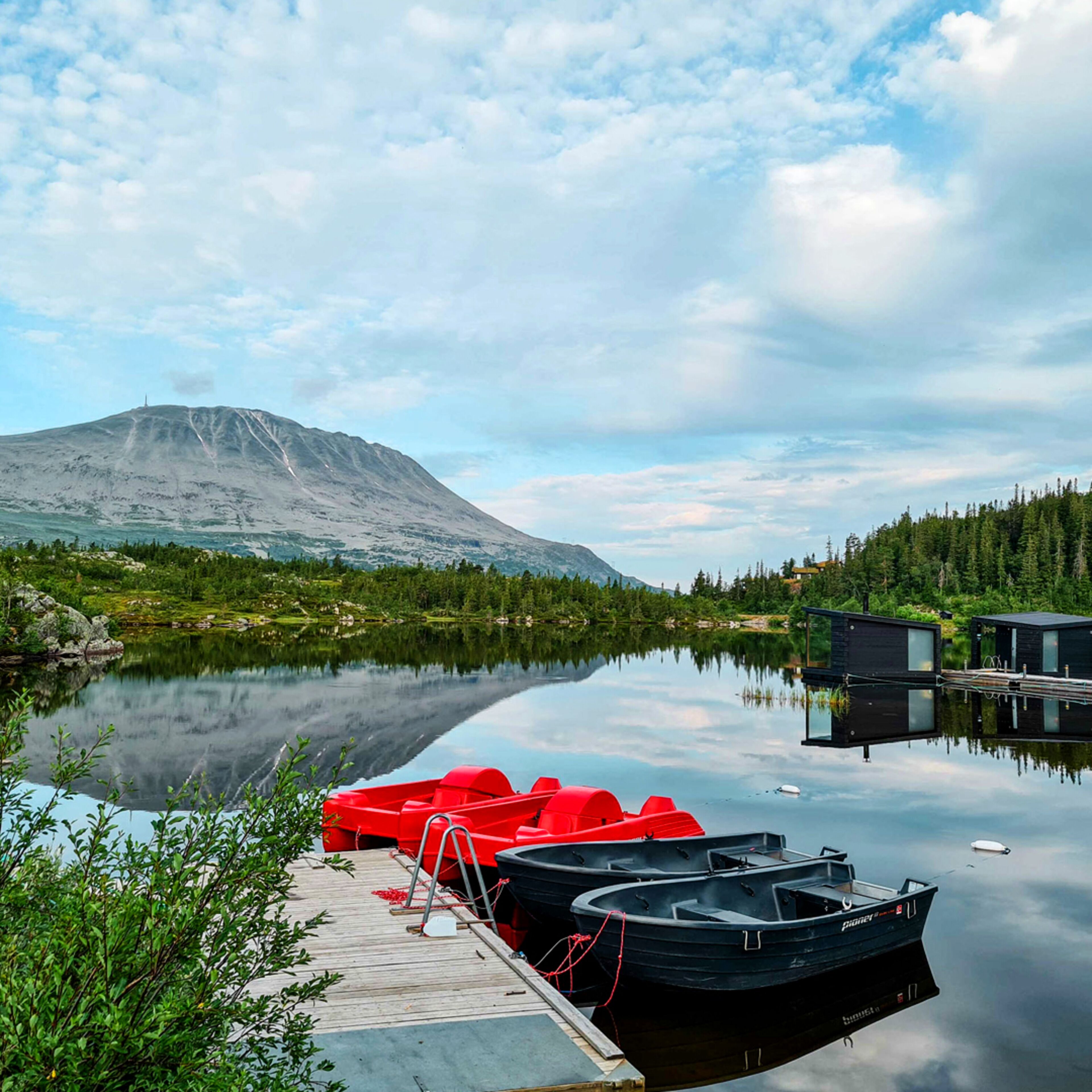 A calm lake with boats docked at a wooden pier, surrounded by greenery and a mountain in the background.