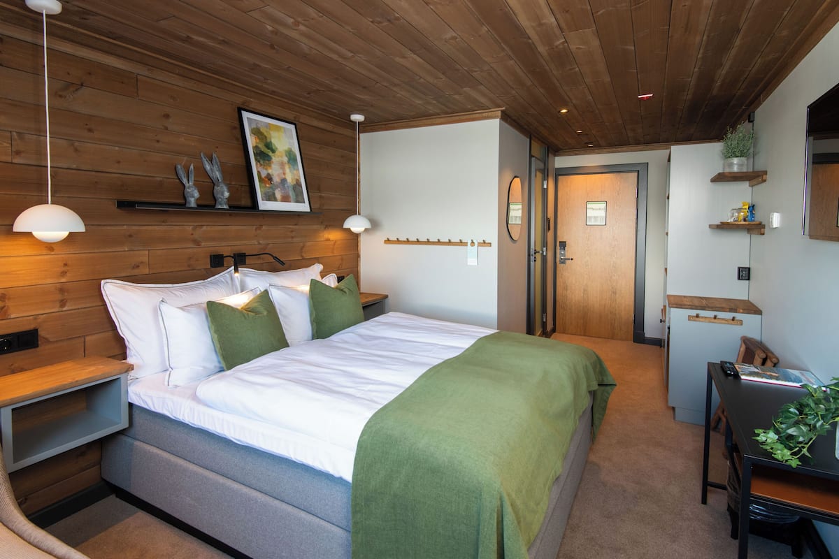 A modern bedroom with a wooden ceiling, a bed with white linens and green pillows, a bedside table, a desk with a chair, and a small plant.