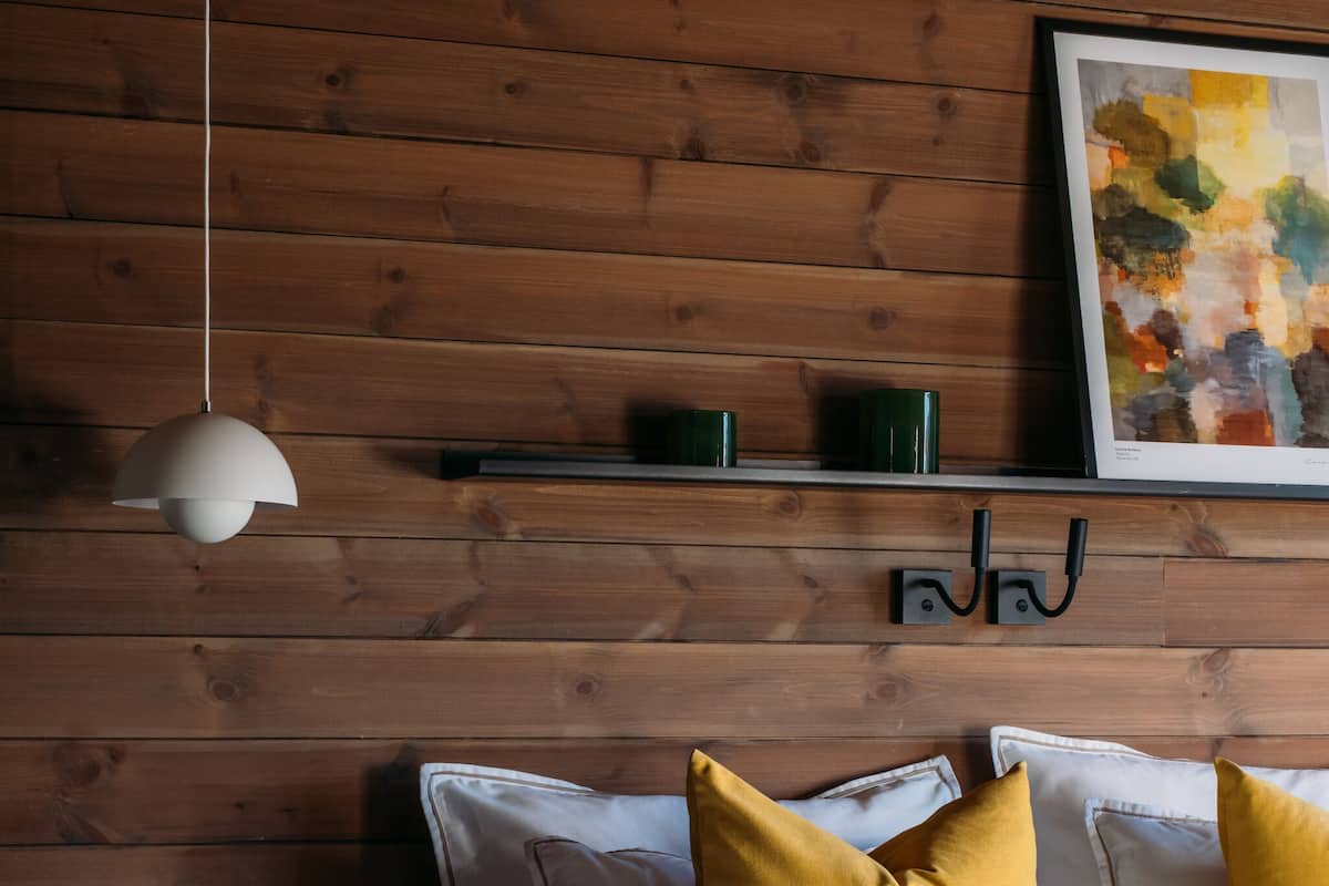 A modern bedroom with a wooden wall, a bed with white and mustard pillows, a pendant light, and a framed abstract painting.