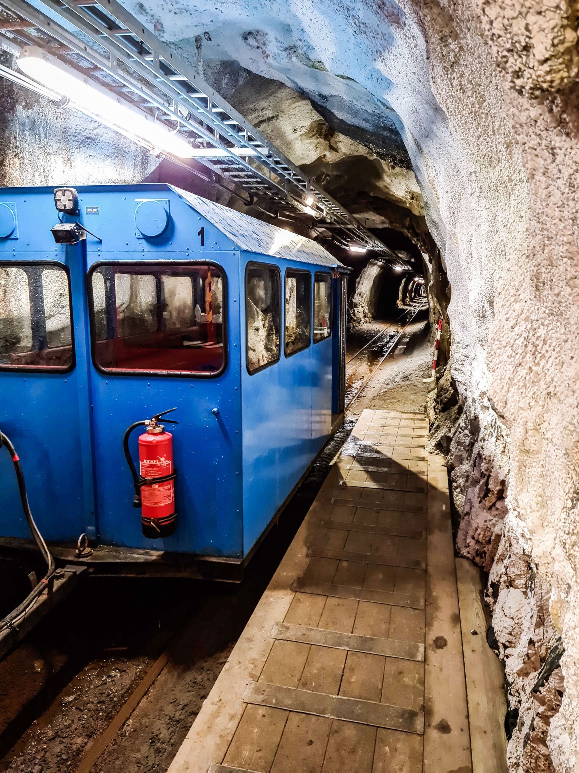 Un wagon de train bleu, équipé d’un extincteur et de matériel de sécurité, traverse un tunnel doté d’un quai en bois.