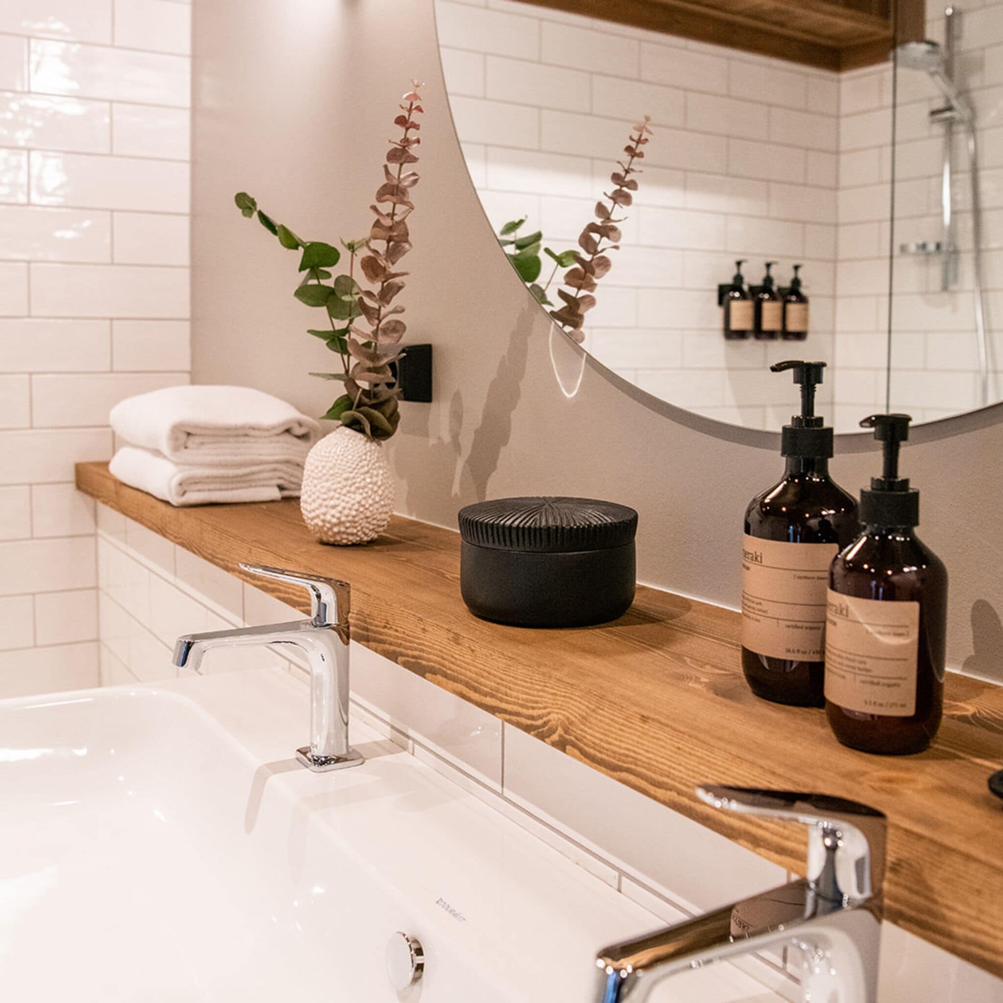 A modern bathroom with a white sink, wooden countertop, and a mirror. There are two bottles of shampoo and a black container on the counter.