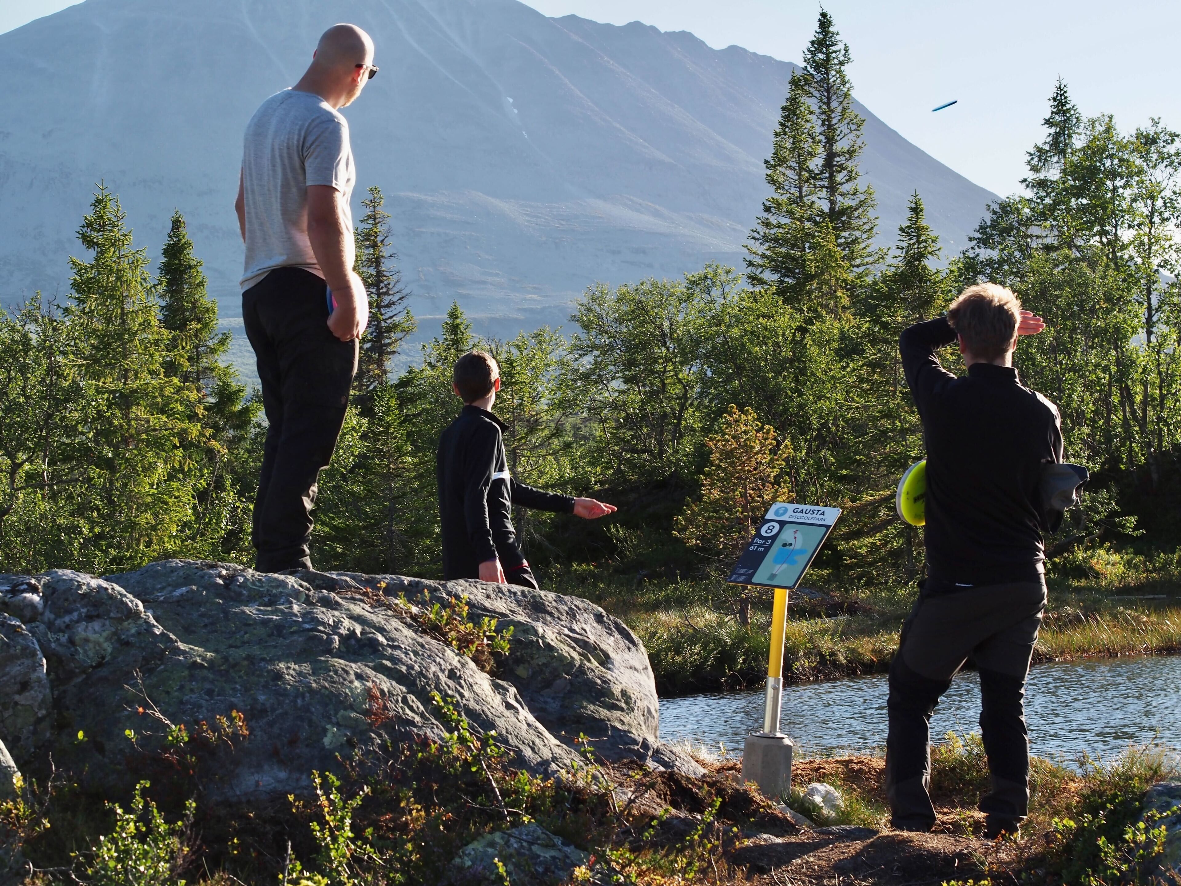 A group of people standing by a lake with a mountain in the background and a sign indicating a location.