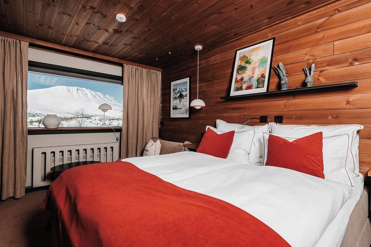 A bedroom with a large bed, red and white bedding, wooden walls, a window with a view of a mountain, and two wall-mounted artworks.