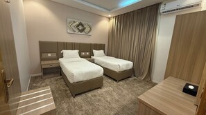 Habitación de hotel con dos camas, una mesita de noche, una lámpara de noche, una obra de arte colgada en la pared, una ventana con cortinas y un equipo de aire acondicionado.