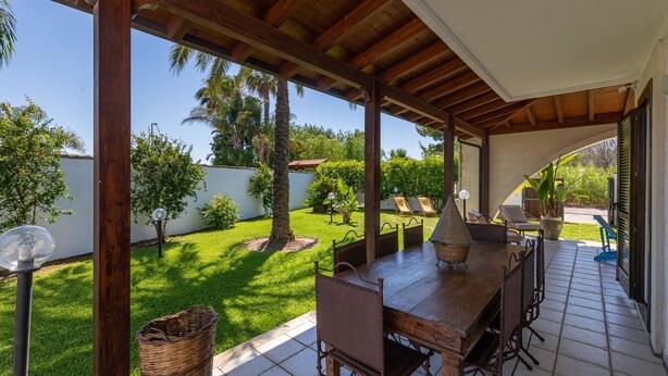 Eine überdachte Terrasse mit Holztisch und -stühlen, umgeben von üppigem Garten und Palmen.
