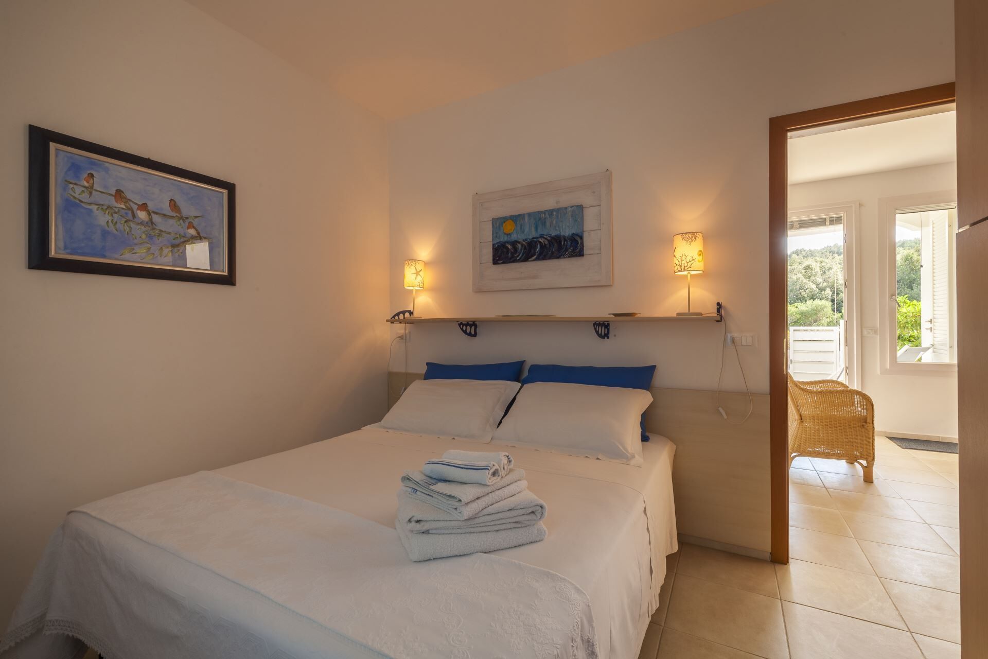 A bedroom with a bed, a framed painting, two wall-mounted lamps, and a view of the outdoors.