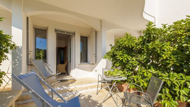 A sunlit patio with a stone floor, a table with chairs, and a large green bush. - Casa Bellaria | RivedelSalento - Casa Bellaria piano terra (Castro)