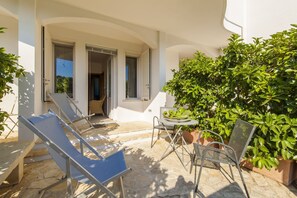 Une terrasse ensoleillée avec un sol en pierre, une table entourée de chaises et un grand arbuste vert.