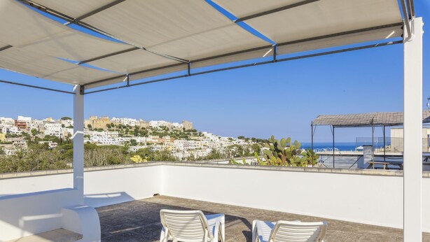 A rooftop terrace with two white plastic chairs, a covered area with a metal frame, and a view of a coastal town with buildings and a clear blue sky. - Casa Bellaria primo piano (Castro)