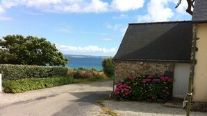A gravel driveway leading to a stone building with a sloped roof, surrounded by hedges and flowering plants.