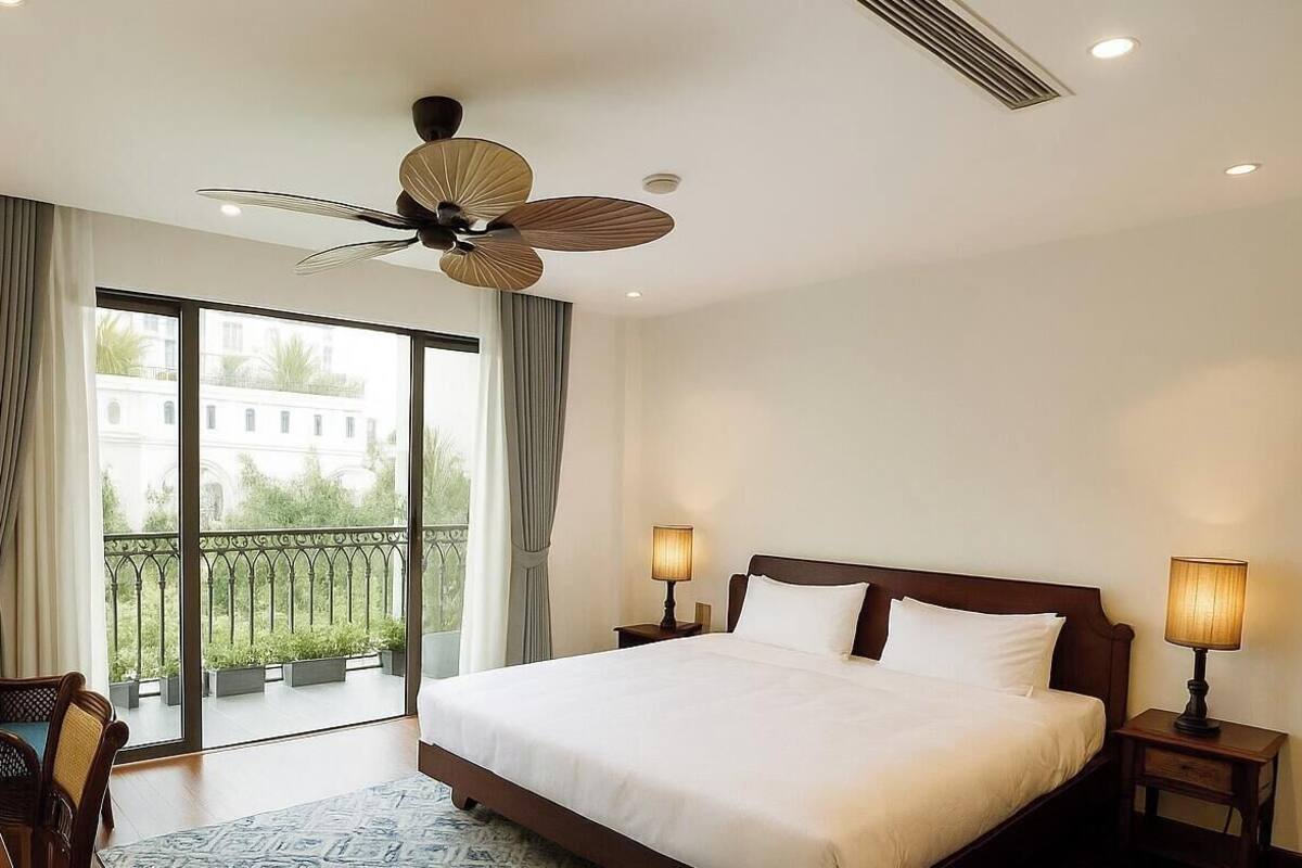 A hotel room with a large bed, a desk with a vase of flowers, a balcony with a view, and a ceiling fan.