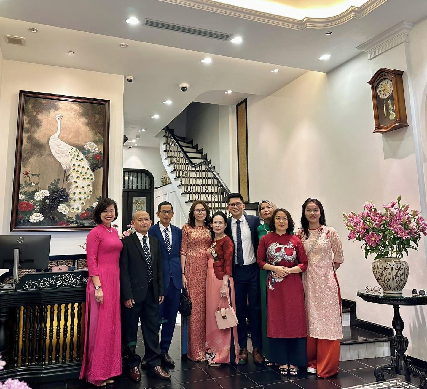 Un grupo de personas posando en el vestíbulo de un hotel, con una escalera, un reloj y un cuadro de un pavo real.