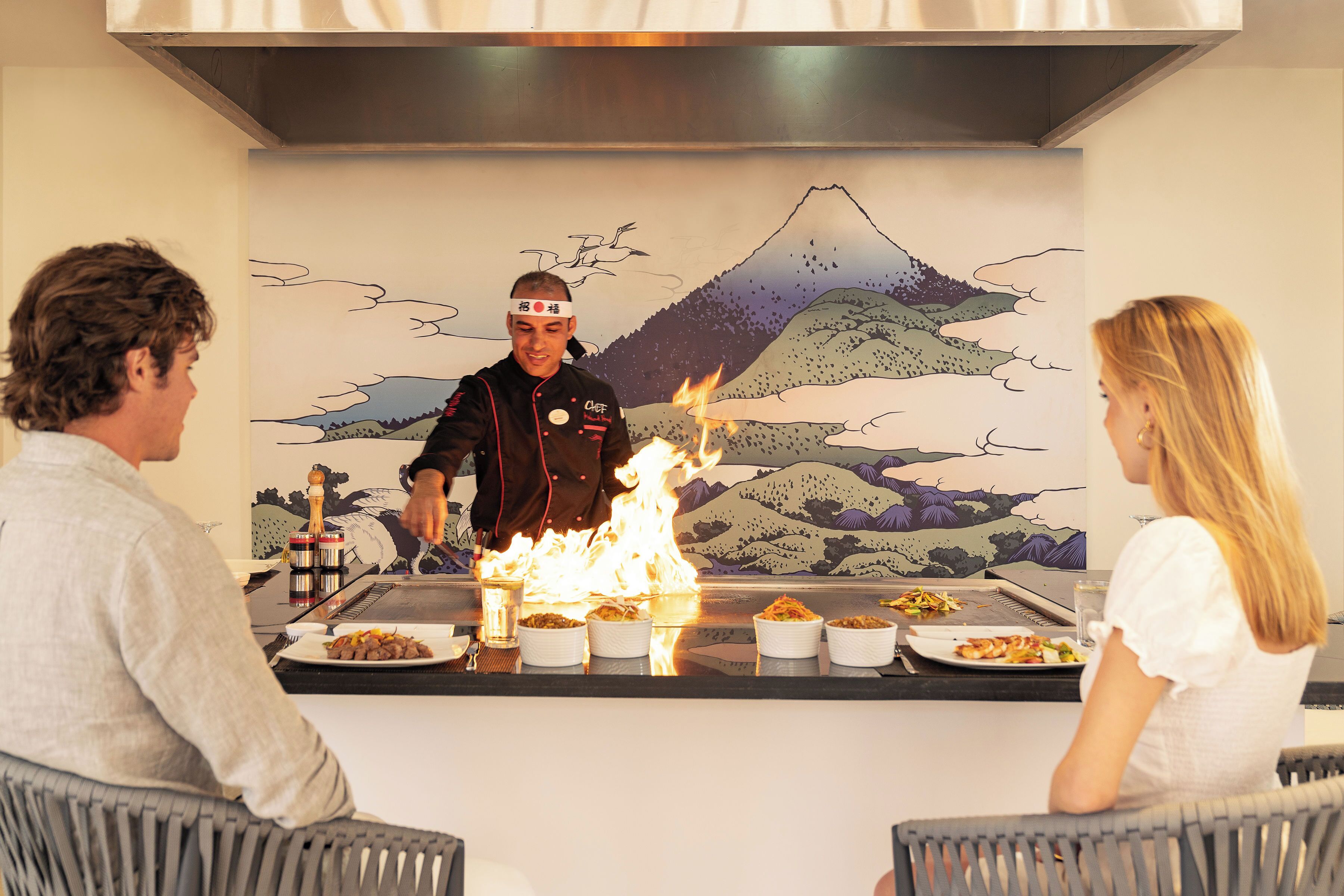 Un restaurante con un chef realizando una técnica culinaria a base de fuego, sirviendo comida a los comensales.