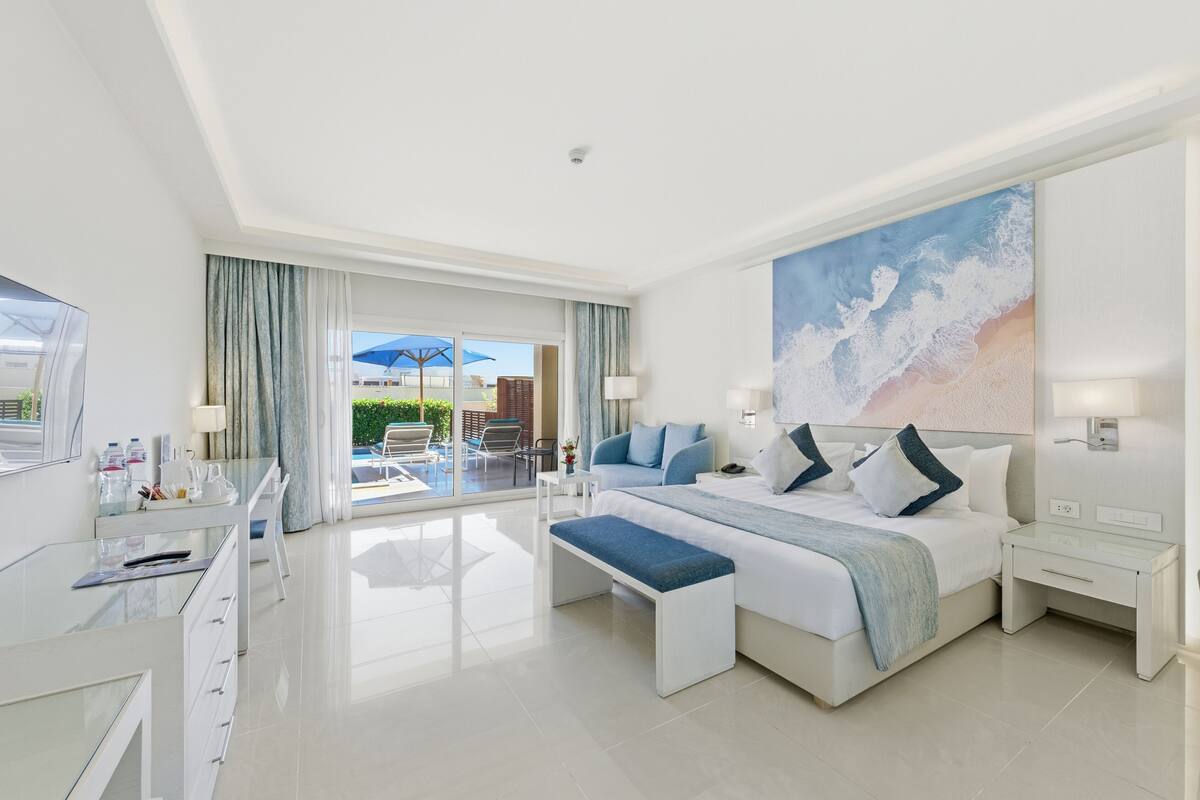 Une chambre d’hôtel moderne avec un grand lit, un bureau en verre et une vue sur l’espace extérieur de la piscine.