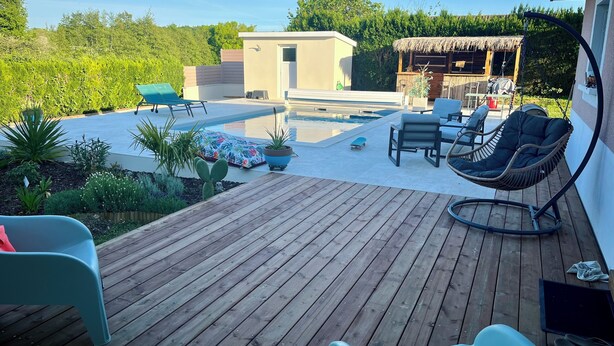 Una terraza de madera con mobiliario exterior, una piscina y una estructura con techo de paja.
