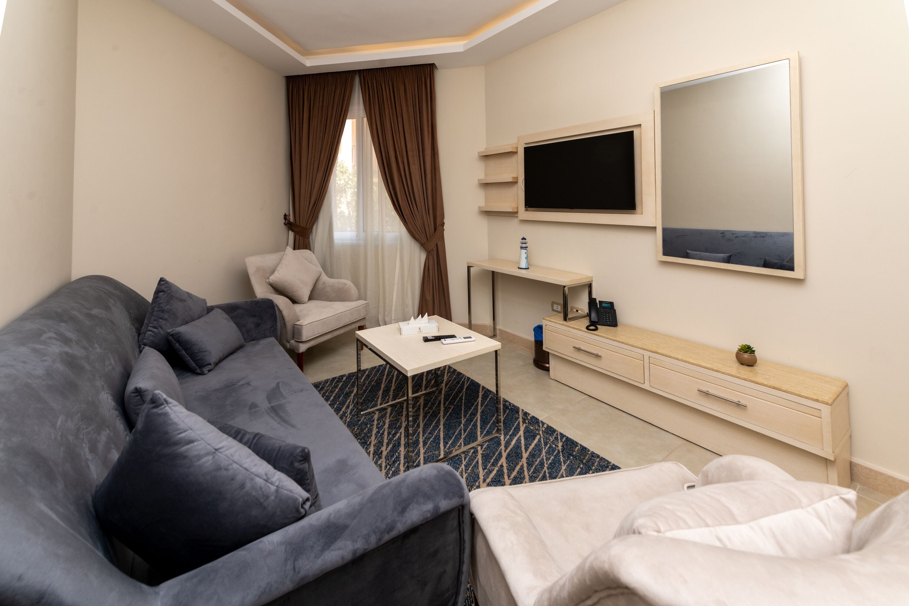 A modern hotel room with a grey sofa, a small white table, a TV, and a wooden cabinet.