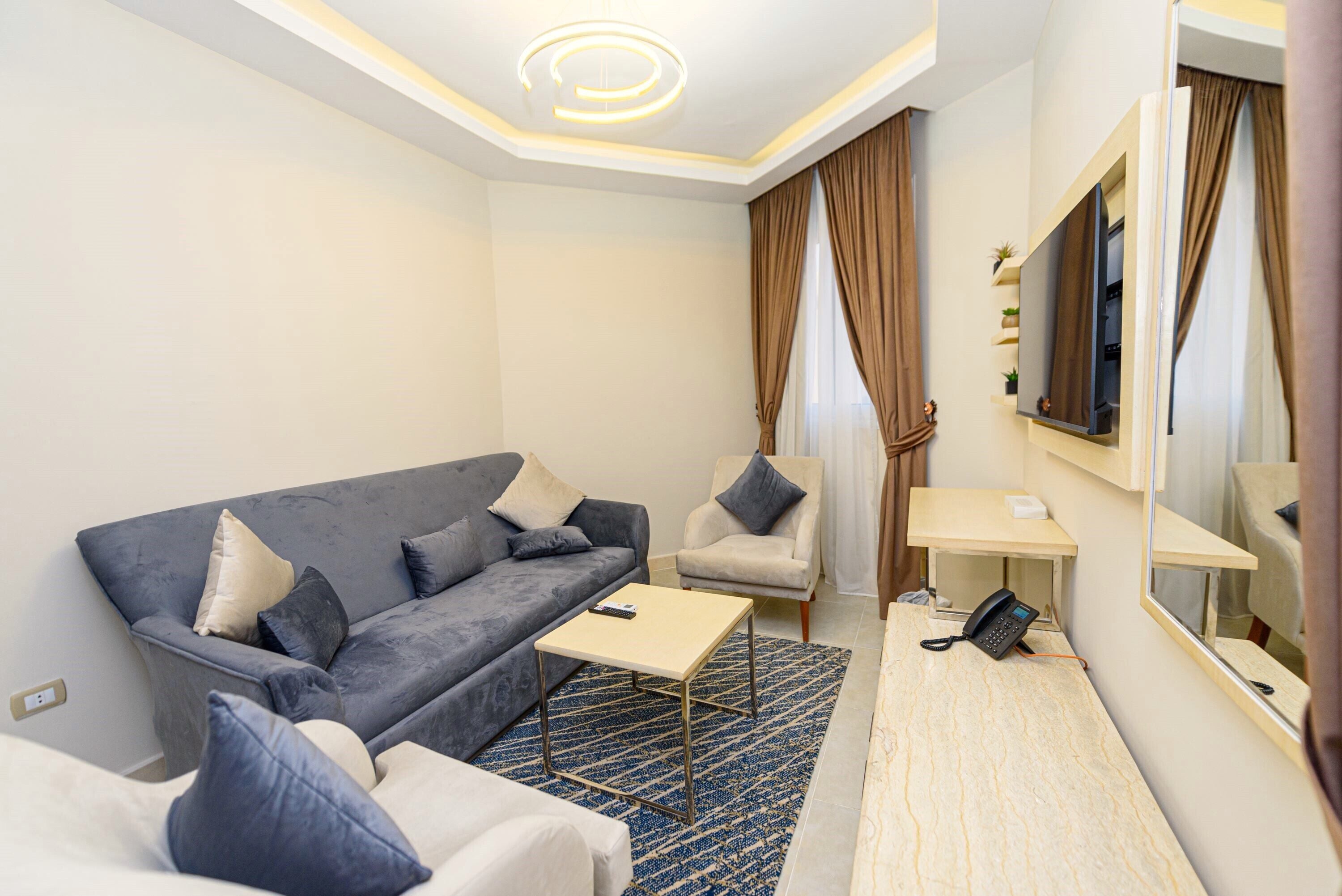 Una habitación de hotel moderna con un sofá gris, una mesita pequeña, una silla, una mesita de noche con un teléfono y un espejo grande.