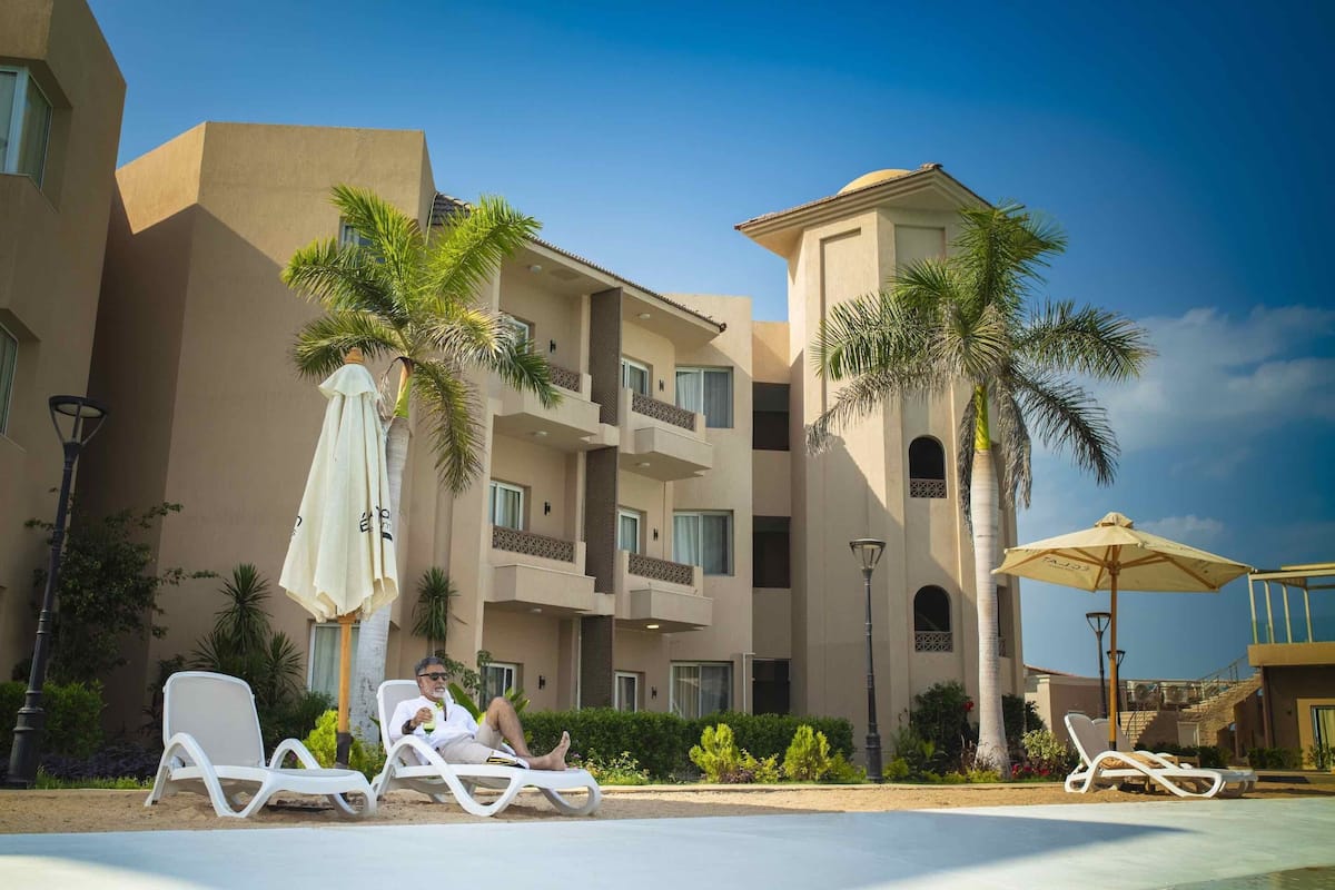 Un hotel con varios balcones, palmeras y sillones de descanso junto a la piscina.