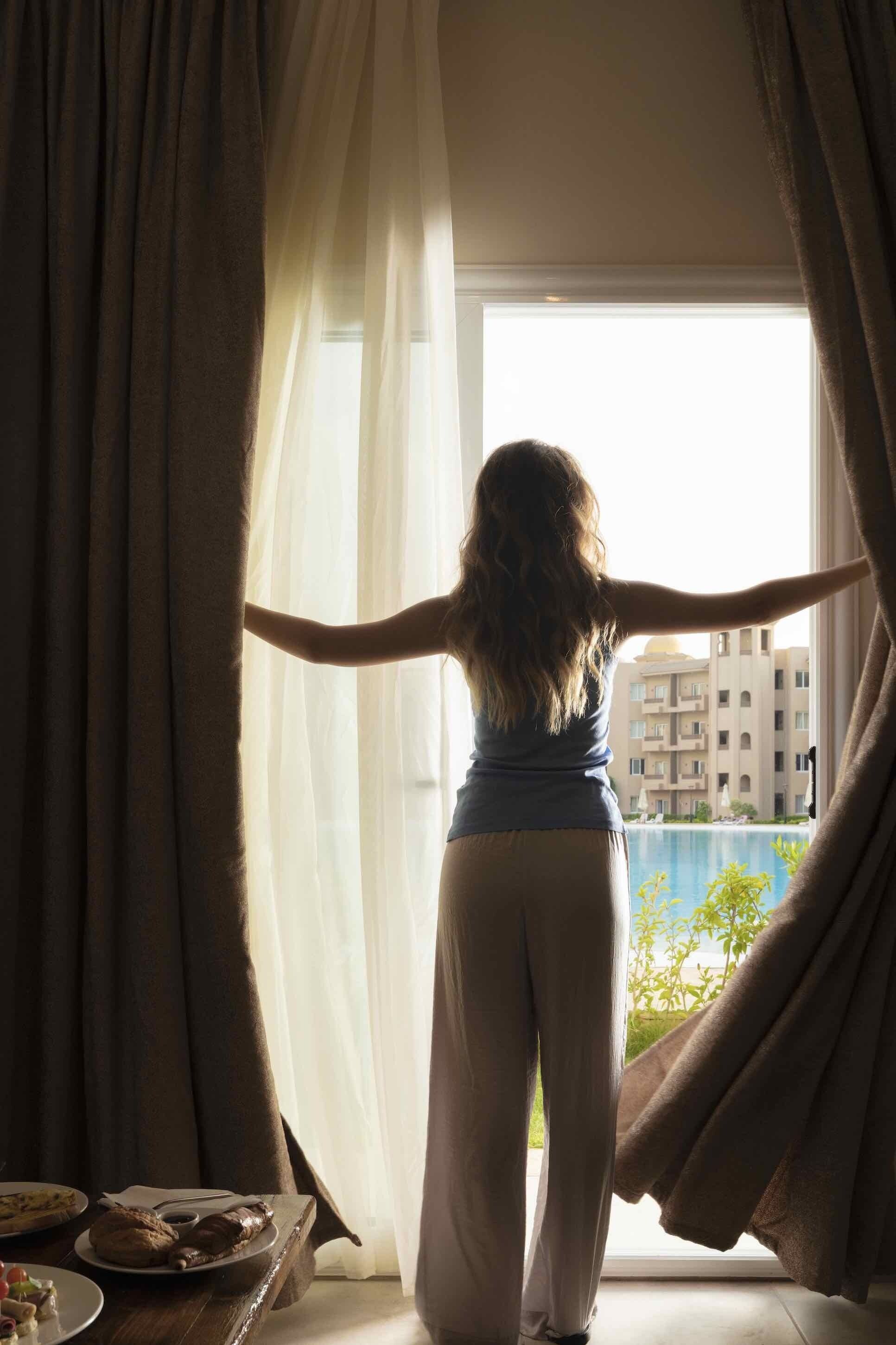 A person standing by a window with curtains, facing a view of a building and a pool.