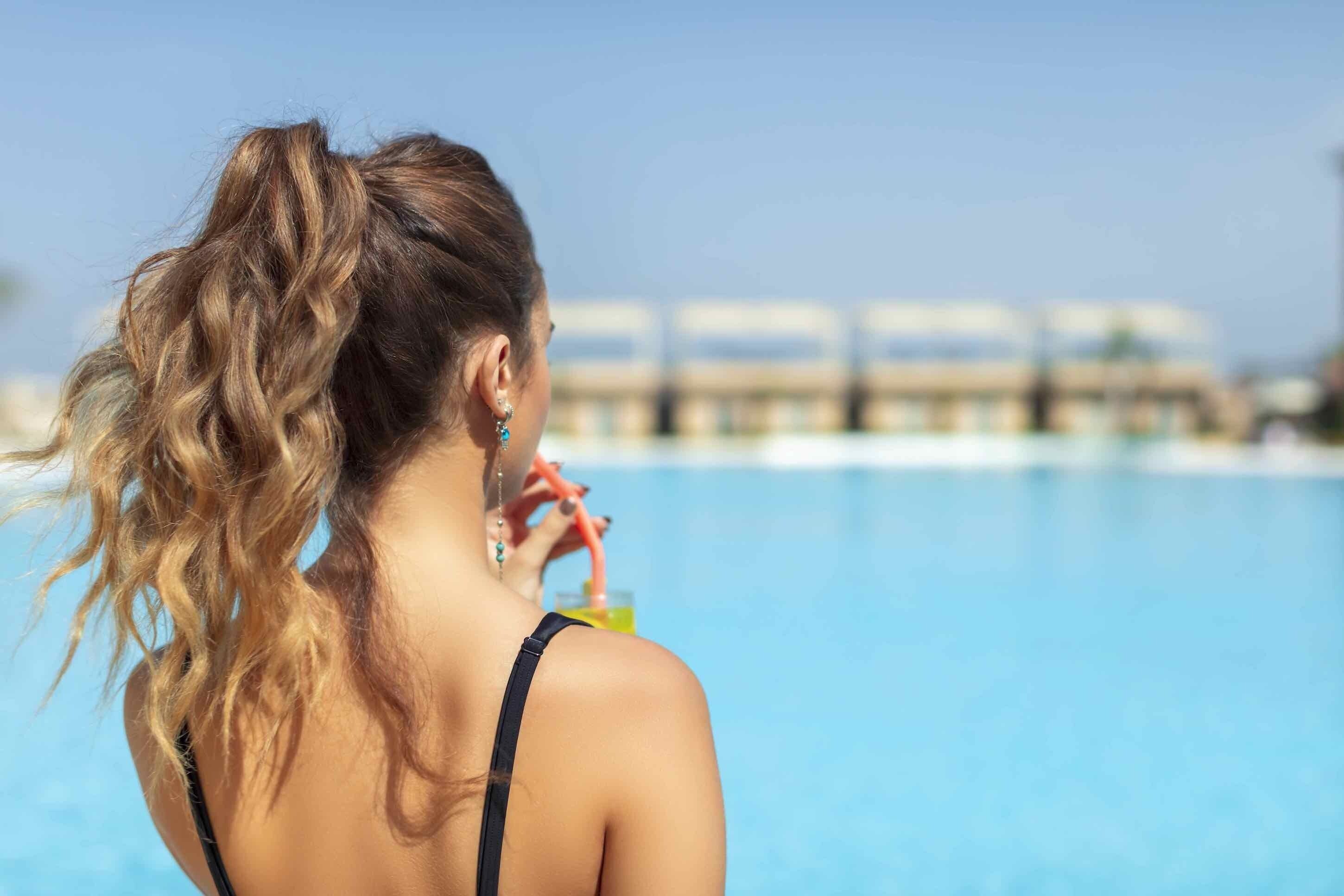 Une personne, les cheveux attachés en queue de cheval, se trouve près d’une piscine, d’où l’on a une vue dégagée sur l’eau et sur les bâtiments situés au loin.