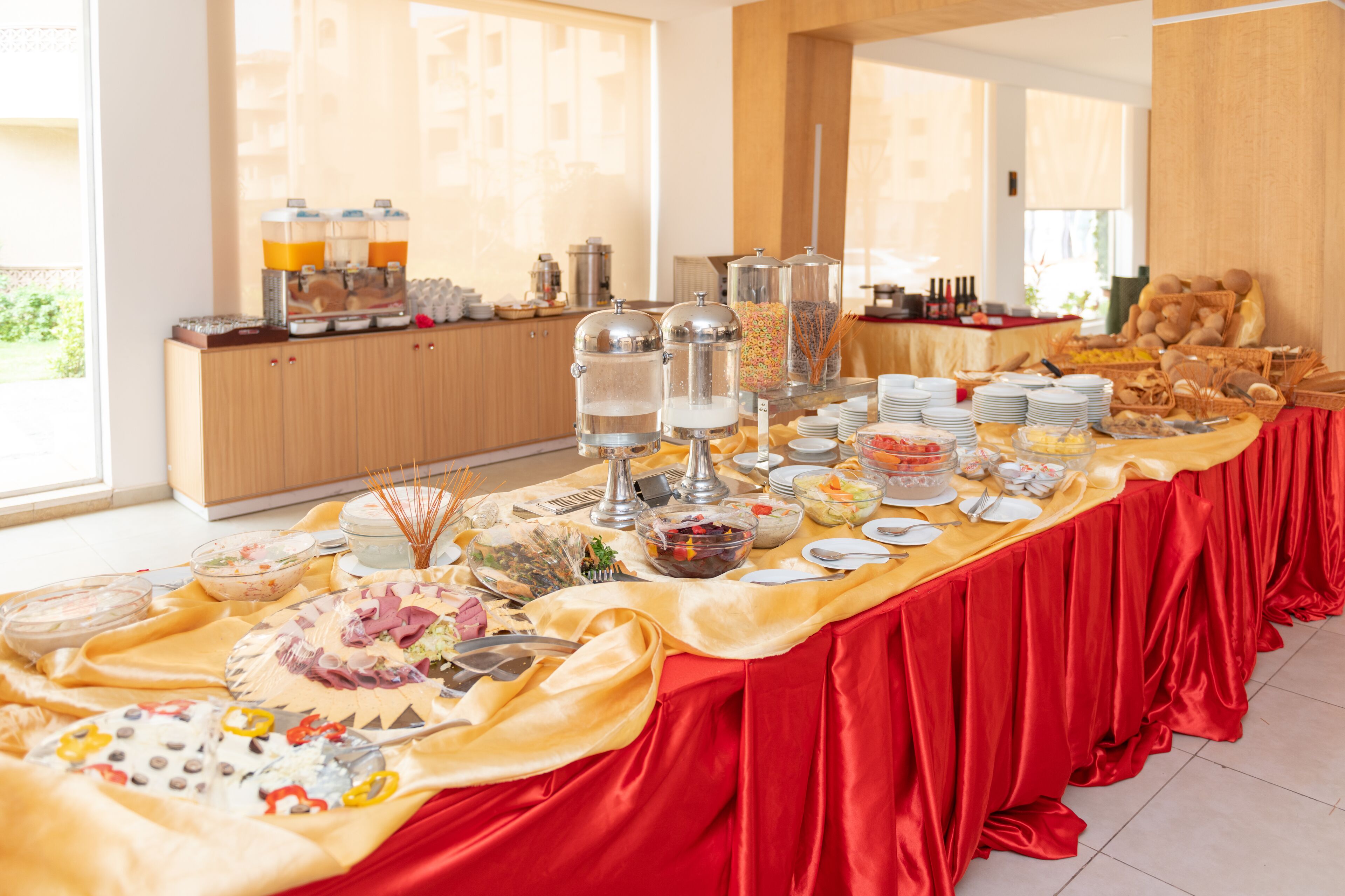 A buffet table with various food items and beverages, set in a bright room with large windows.