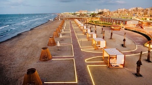 Ein Hotel direkt am Strand mit einer langen Reihe von Liegestühlen und Sonnenschirmen, einem Weg zum Strand und im Hintergrund Meer und Stadtlandschaft.