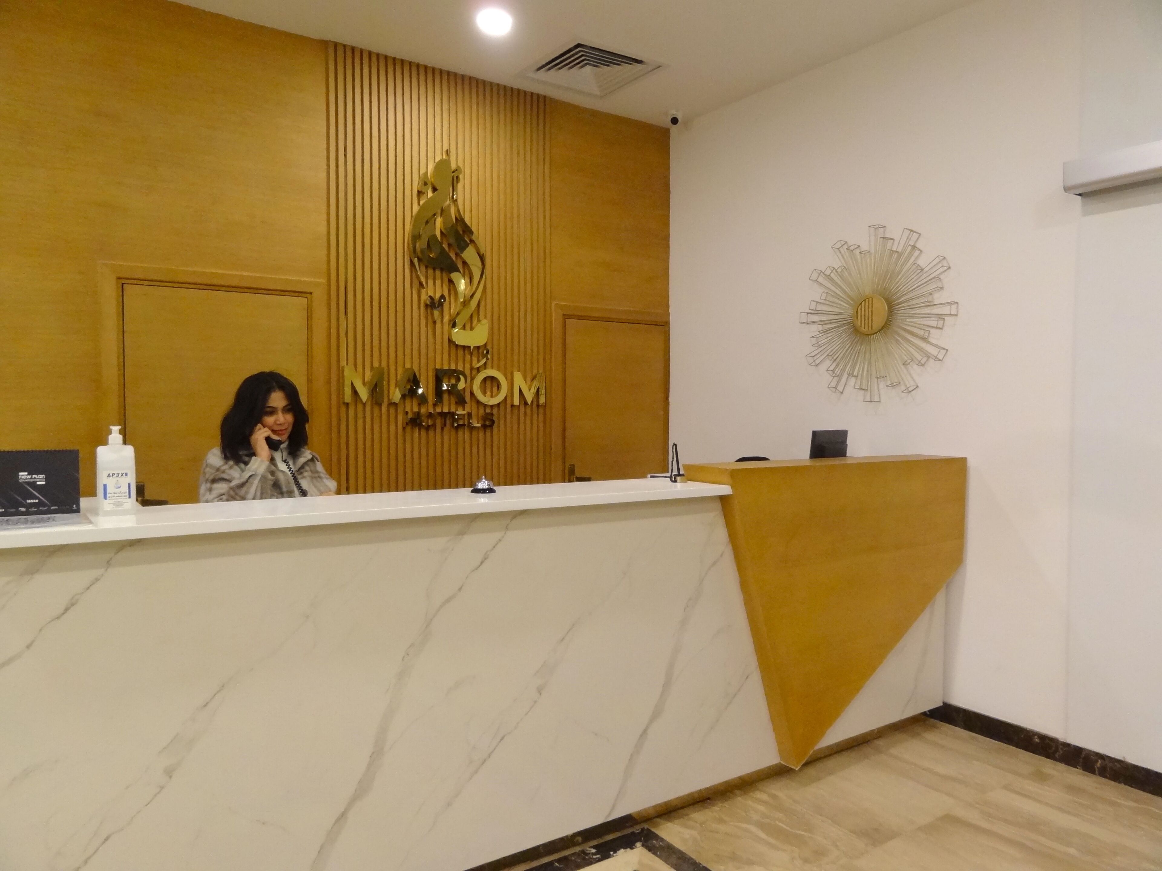 Una recepción con mostrador de mármol, pared revestida de madera con logotipo dorado, un reloj de pared moderno y un dispensador de gel hidroalcohólico en la encimera.