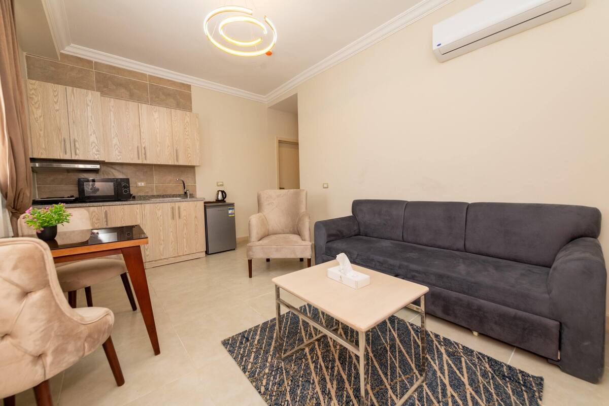 Una sala moderna con un sofá gris oscuro, una mesita con una caja de pañuelos, un sillón beige y una zona de cocina con armarios de madera y un microondas.