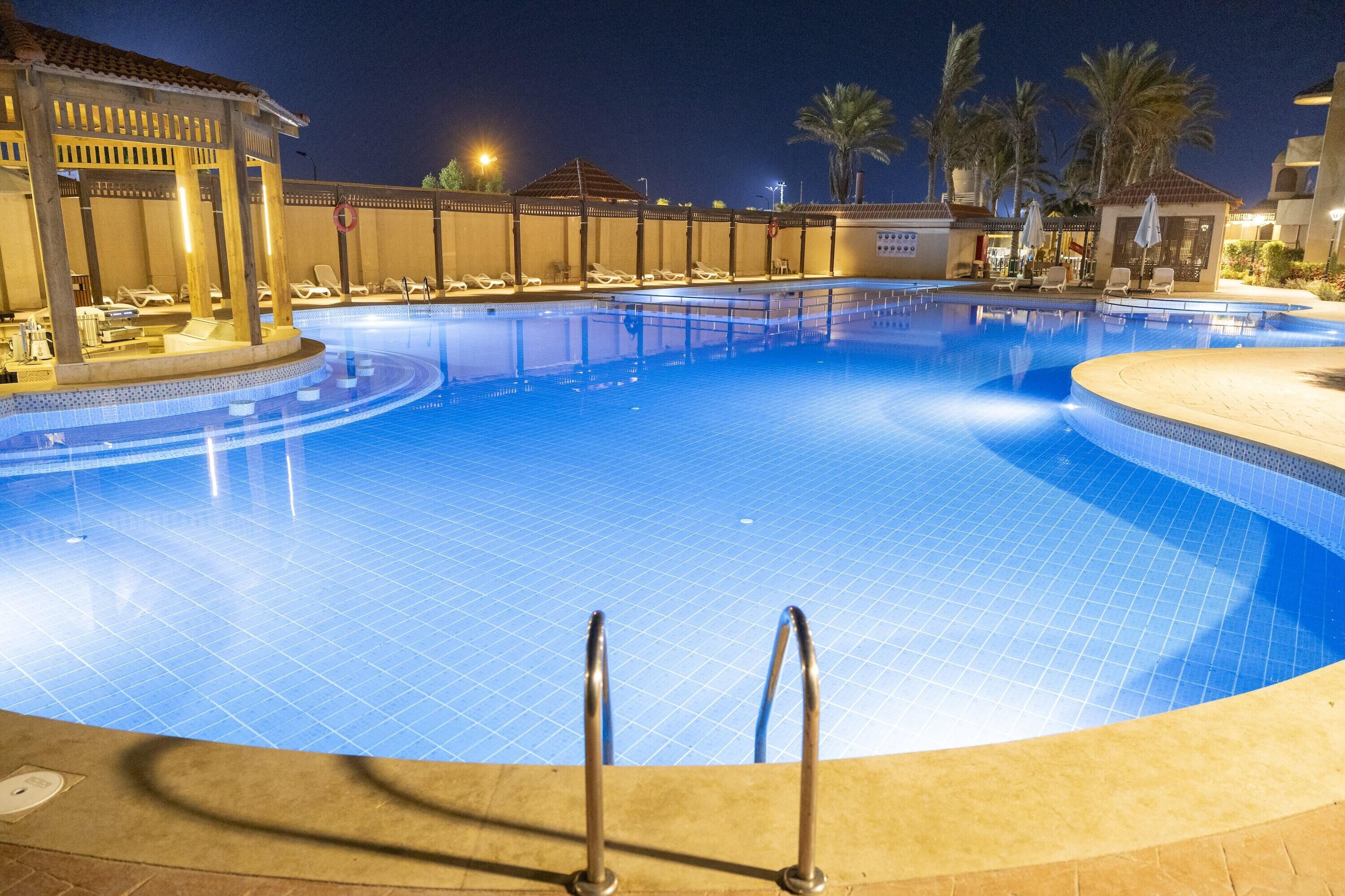 A large, well-lit swimming pool with lounge chairs and a covered area, surrounded by palm trees and a building with a tiled roof.