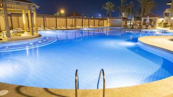A large, well-lit swimming pool with lounge chairs and a covered area, surrounded by palm trees and a building with a tiled roof.